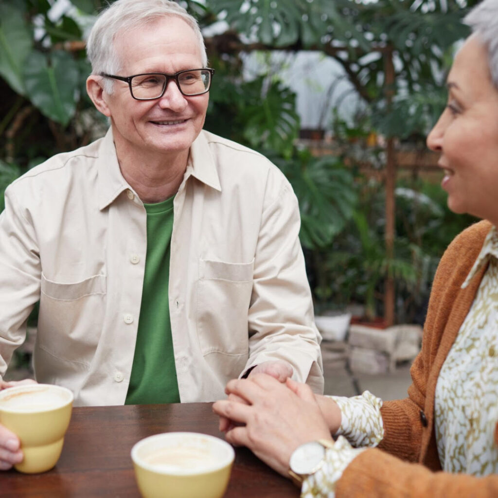 Photo of a gray haired man and woman sitting amidst green plants and drinking coffee. 